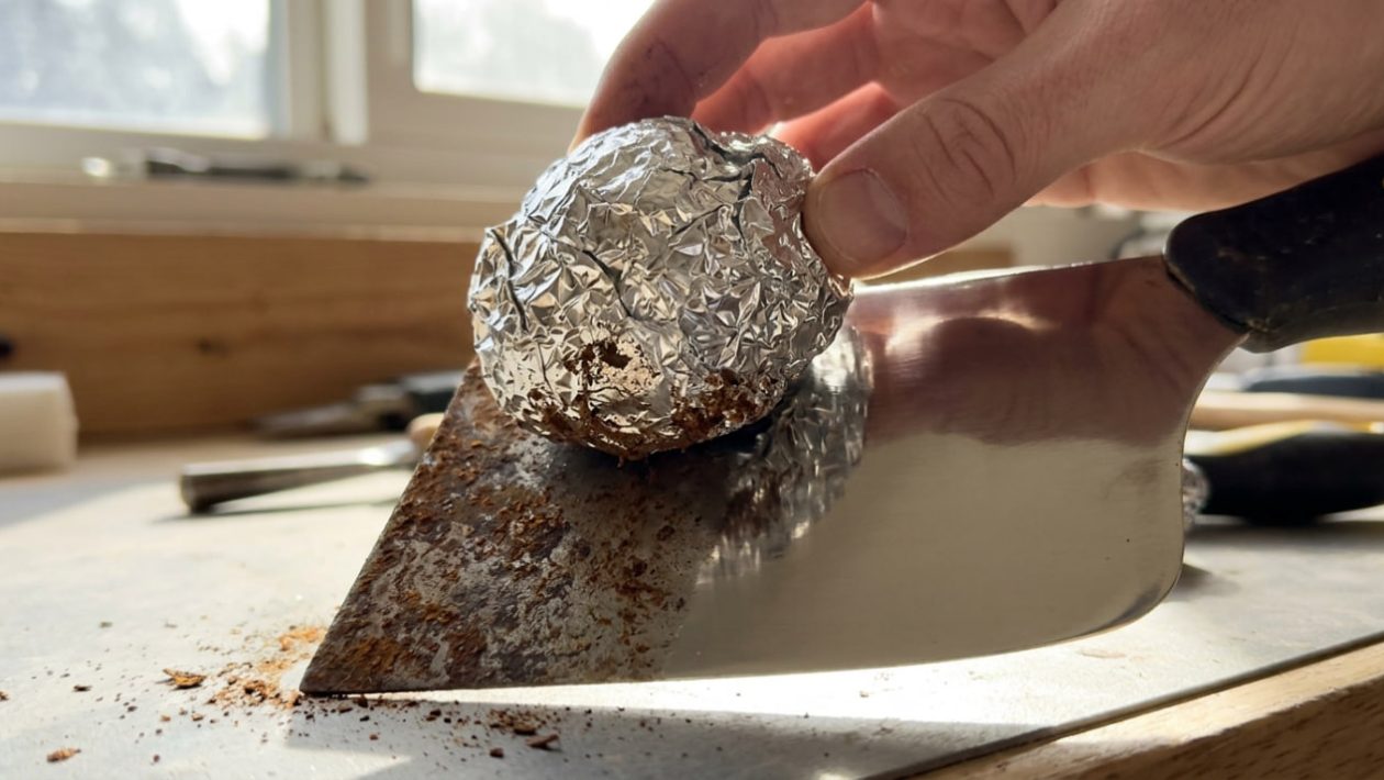 Eine Kugel aus Alufolie rubbelt Rost von einem Spachtel und hinterlässt glänzendes Metall.